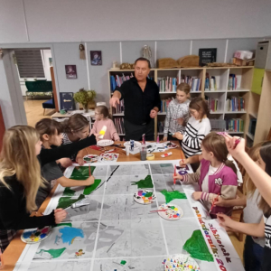 Na zdjęciu dzieci z prowadzącym warsztaty pod hasłem "do dzieła", przed nimi na stole ułożona przez nich mapa Torzymia, którą malują.zaznaczjąc swoje miejsca zamieszkania.