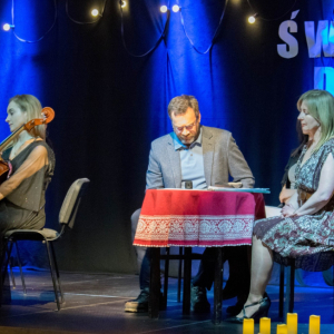 Na pierwszym planie aktorzy, jeden z nich czyta w utwór po lewej stronie siedzi zasłuchana wiolonczelistka. 