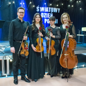 Członkowie Urban String Quartet stoją przed sceną z instrumentami, uśmiechnięci i wyraźnie zadowoleni po występie. Od lewej: pierwszy skrzypek Mateusz Makuch, skrzypaczka Weronika Podklu, altowiolistka Natalia Rychlik oraz wiolonczelistka Natalia Makuch. Każde z nich trzyma w dłoni czerwoną różę – symbol uznania i podziękowania za wyjątkowy koncert.