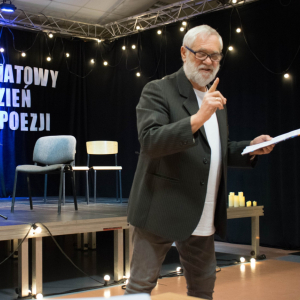 Pan Krzysztof Stróżański podczas wykładu poświęconego twórczości Andrzeja Bursy. Elegancko ubrany w czarną marynarkę, z uśmiechem na twarzy prowadzi spotkanie, unosząc wskazujący palec prawej ręki, podkreślając ważny fragment wypowiedzi. W lewej dłoni trzyma notatki, które towarzyszą jego barwnej i angażującej narracji.