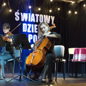 Na pierwszym planie widoczna jest wiolonczelistka oraz towarzysząca jej altowiolistka – obie artystki w pełnym skupieniu oddają się grze. Ich zaangażowanie i koncentracja podkreślają wyjątkowy, kameralny charakter występu. Scena utrzymana jest w ciemnej tonacji – czarne tło rozświetlają subtelne lampki oraz delikatne, niebieskie światło, które nadaje całości nastrojowy i elegancki klimat.