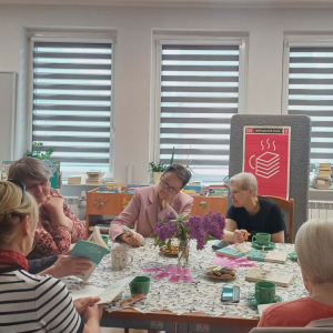 Na zdjęciu widać siedem naszych klubowiczek przy stole, na którym nie brakuje kawy i ciasteczek – nieodłącznego elementu spotkań Dyskusyjnego Klubu Książki. Panie są pochłonięte rozmową i wspólnym spędzaniem czasu.