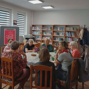 Na zdjęciu widać siedem naszych klubowiczek przy stole, na którym nie brakuje kawy i ciasteczek – nieodłącznego elementu spotkań Dyskusyjnego Klubu Książki. Panie są pochłonięte rozmową i wspólnym spędzaniem czasu.