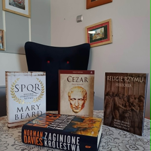 Na stoliku stoją trzy książki od lewej:  M. Beard "SPQR. Historia starożytnego Rzymu", A. Goldsworthy "Cezar> Życie giganta", M. Beard, J. North,S. Price "Religie Rzymu. Historia".