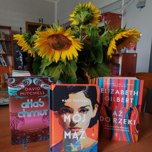 Na zdjęciu trzy książki od prawej: "Aż do rzeki"- Elizabeth Gilbert, "Mój mąż"-Maud Ventura, "Atlas chmur"- Dawid Mitchell. Książki sotjąna stole na tle żółtych słoneczników.
