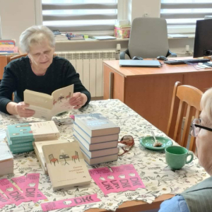 Na zdjęciu: Dwie członkinie Dyskusyjnego Klubu Książki podczas spotkania. Jedna z pań czyta fragment omawianej książki, druga z zainteresowaniem słucha. Na stole widoczne są książki oraz zakładki z logo Dyskusyjnego Klubu Książki.