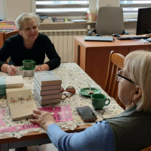 Na zdjęciu: Członkinie Dyskusyjnego Klubu Książki podczas rozmowy o omawianej lekturze. Panie dzielą się swoimi spostrzeżeniami i wrażeniami z przeczytanej książki. Na stole widoczne są książki oraz zakładki z logo Dyskusyjnego Klubu Książki.