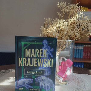 Zdjęcie przedstawia książkę stojąca na stole. Książka autorstwa Marka Krajewskiego o tytule"Drgoa krwi".