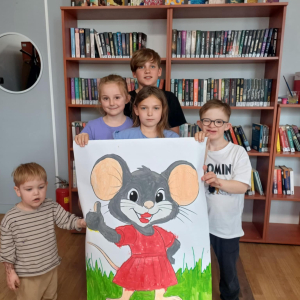 Na zdjeciu 5 dzieci trzymajaca przed sobą skonczony pomalowany przez nich plakat przedstawiuajacy Mysz matyldę. Mysz ma czerwoną sukienkę, jest szara, stoi na zielonej trawie i ma pięknie pomalowane na czerwono usta. 